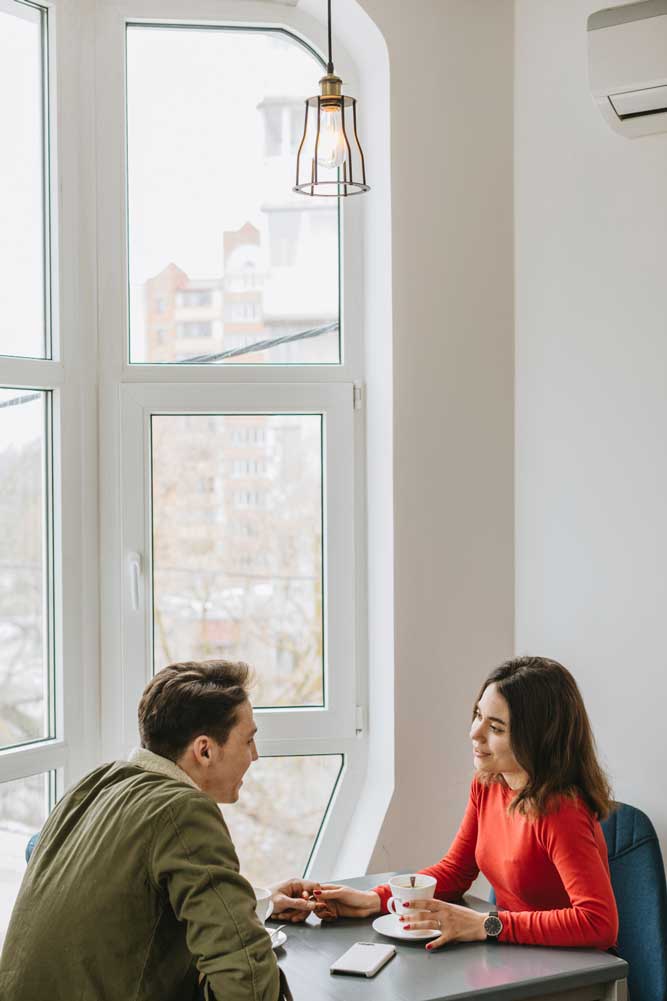 Couple sitting at a table by a large white-framed window enjoying coffee and conversation in a bright modern interior, highlighting natural light, comfort, and professional Window Replacement Atlanta services for stylish, energy-efficient spaces.