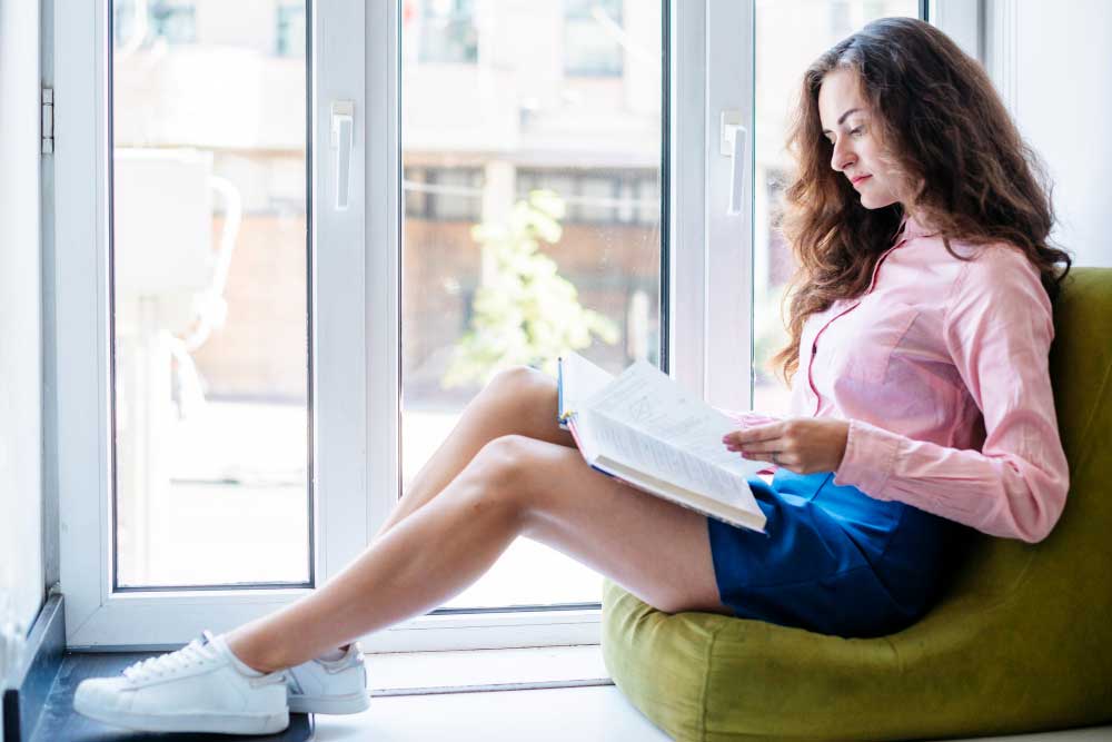 Woman relaxing by large white-framed windows while reading a book in a bright modern interior, enjoying natural light and comfort, showcasing stylish living spaces and professional Window Replacement Atlanta services.