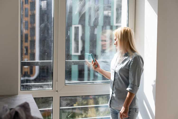Woman standing by a large picture window in a bright apartment, enjoying natural light and city views while using her phone, showcasing modern design and professionally installed Picture Windows Atlanta for clear views and energy efficiency.
