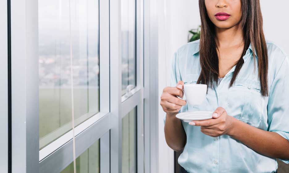 Woman holding a cup of coffee while standing beside large picture windows in a bright modern interior, enjoying natural light and outdoor views, showcasing comfort, style, and professionally installed Picture Windows Atlanta.